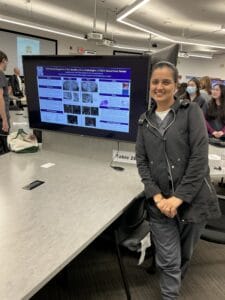 A woman stands and smiles in front of a scientific poster presentation displayed on a large monitor in a classroom or conference setting.