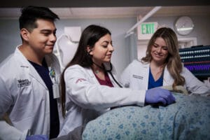 Three medical professionals in white coats examine a patient using a stethoscope and monitor vital signs in a clinical setting.