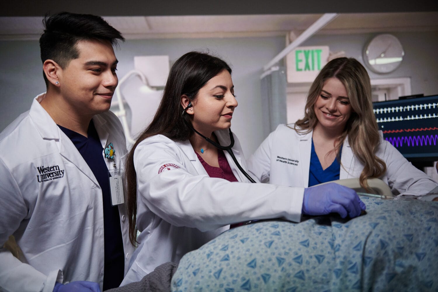 Three medical professionals in white coats examine a patient using a stethoscope and monitor vital signs in a clinical setting.