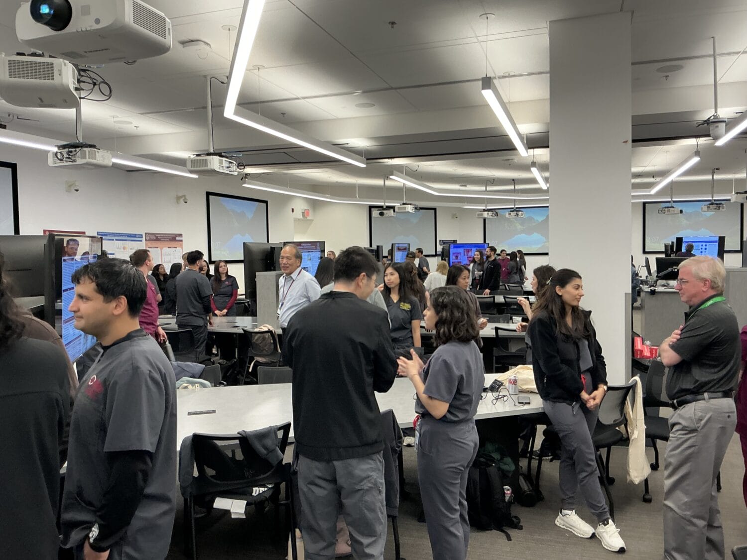 People in scrubs stand and talk in a large, modern classroom with several tables, monitors, and ceiling-mounted projectors.
