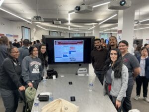 A group of people pose around a table displaying a research poster on a large screen in a conference room, with other attendees and posters visible in the background.