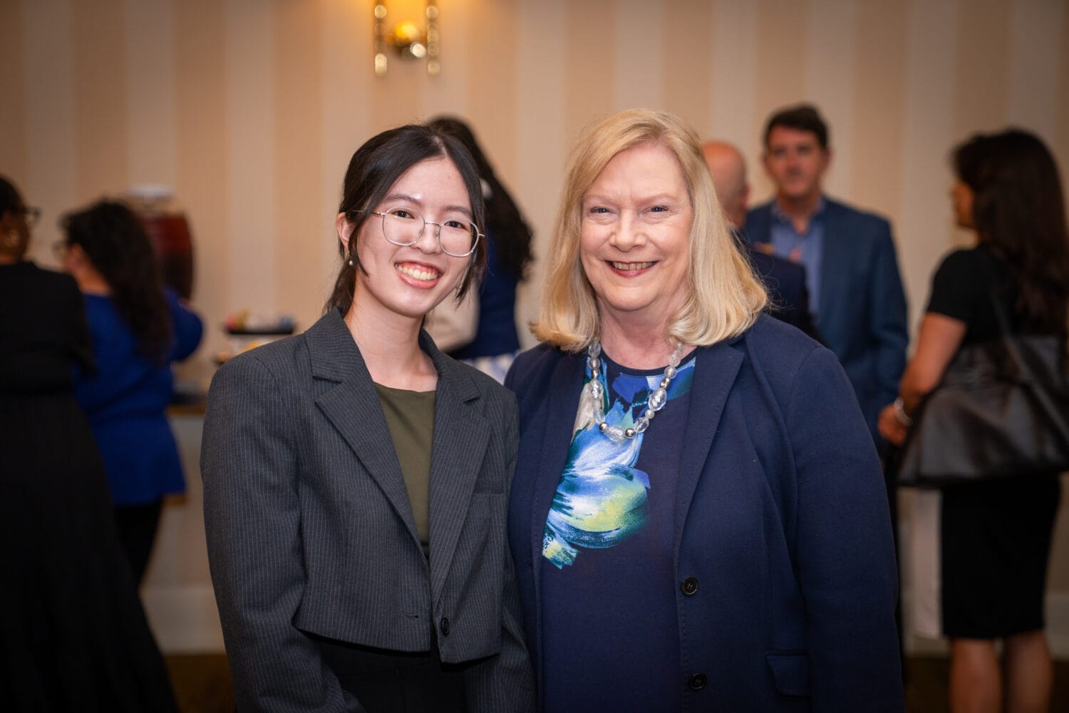 Two women in business attire stand side by side, smiling at the camera in a room with several people and refreshments in the background.