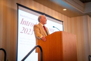 A man in a tan suit speaks at a podium in front of a projected presentation titled "Introducing our 2023" at an indoor event.