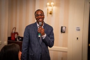 A man in a suit smiles while speaking into a microphone in a conference room with striped wallpaper and wall sconces.