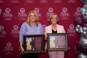 Two women stand in front of a Western University College of Dental Medicine backdrop, each holding a framed plaque with a photo and text.