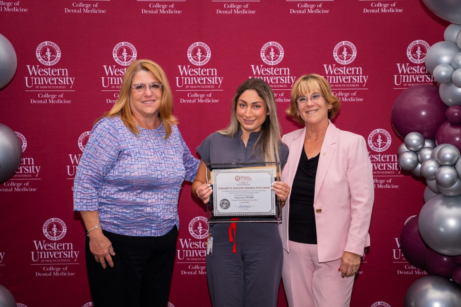 Three women stand in front of a Western University College of Dental Medicine backdrop; the woman in the center holds a certificate, and all are smiling.