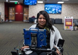A woman in a wheelchair uses a tablet mounted in front of her in a brightly lit indoor setting with office furniture in the background.