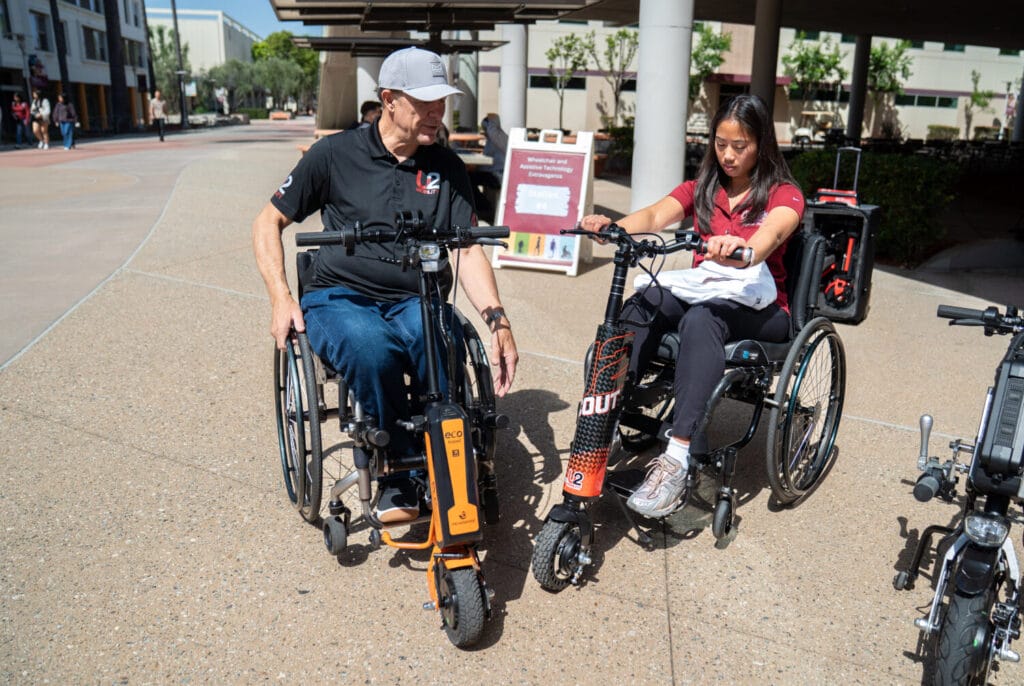 Two people in wheelchairs attach motorized devices to their chairs outdoors on a sunny day.