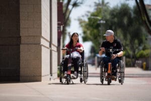Two people using wheelchairs travel along a sidewalk, engaged in conversation on a sunny day near a building and trees.