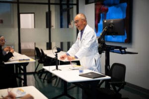 A man in a white lab coat stands at a lectern, speaking to seated students in a modern classroom with a computer monitor and notes on the desks.