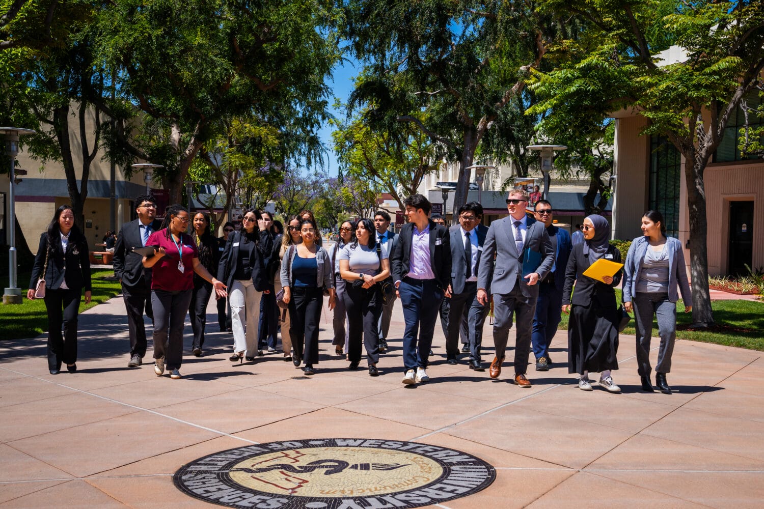 A group of professionally dressed people walks together outdoors on a sunny day, passing a round emblem for Western University of Health Sciences on the pavement.