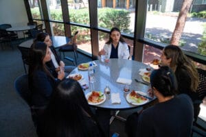 Six people sit around a round table, eating pizza and drinking bottled water, engaged in conversation in a room with large windows.