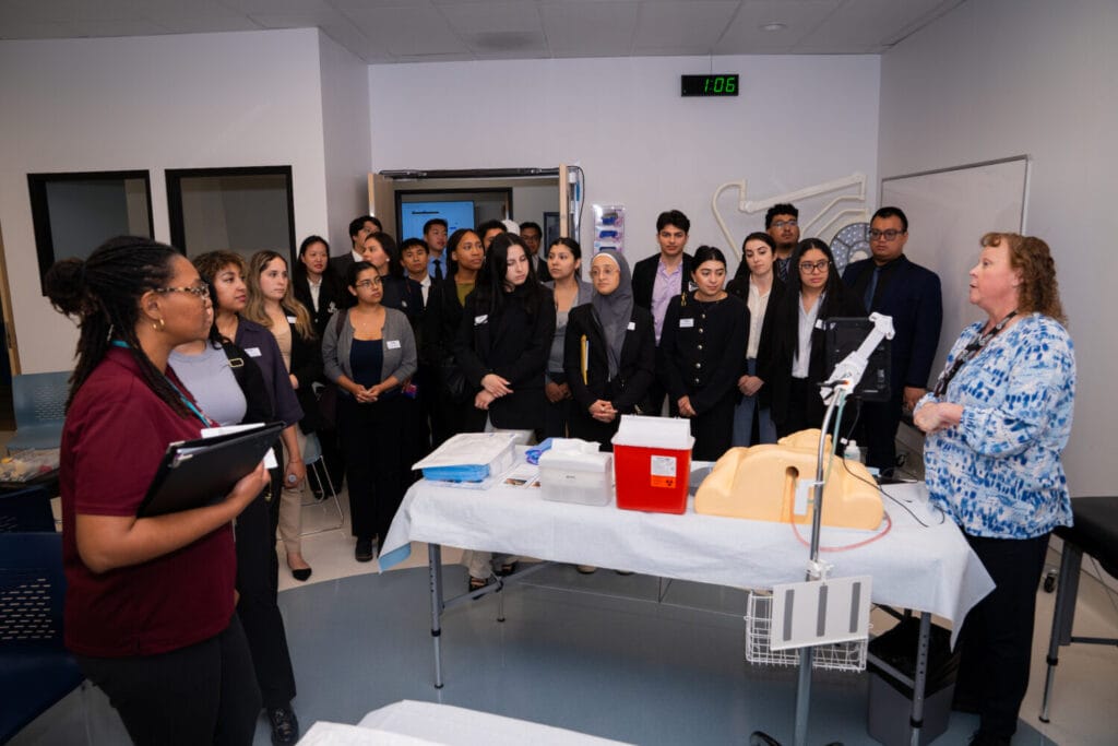 A group of students listens to an instructor in a medical training room with simulation equipment and medical supplies on a table.