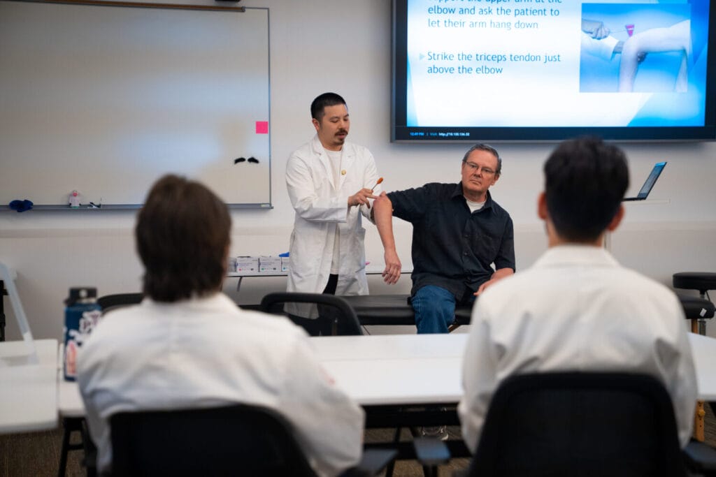 A medical instructor demonstrates an elbow examination on a seated participant while students observe in a classroom with a projected slide on the screen.