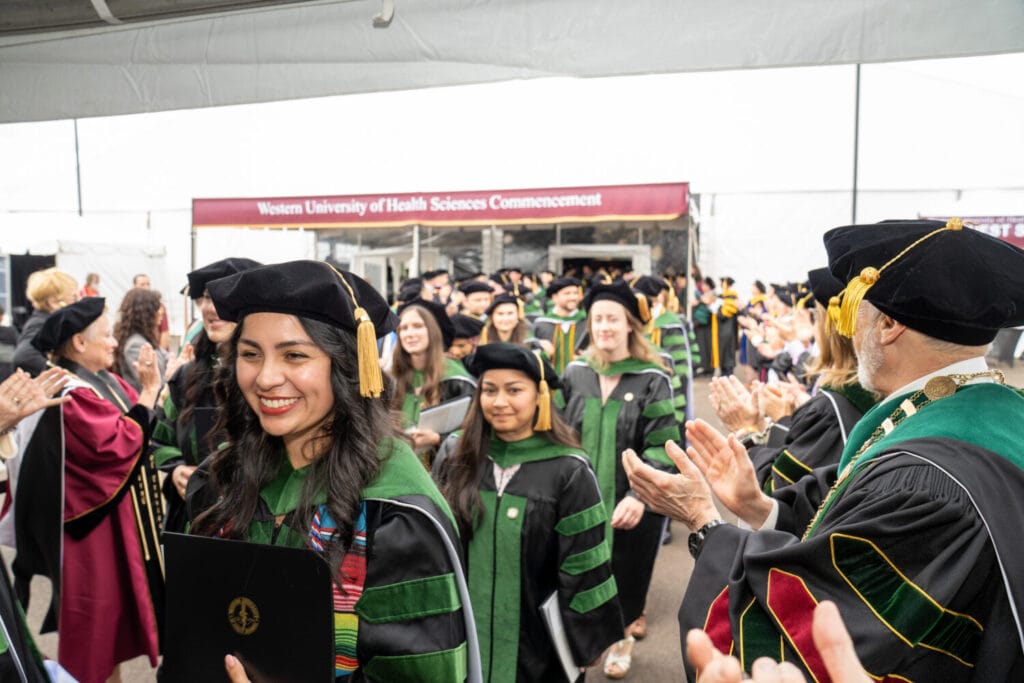 Graduates in academic regalia walk in a procession at a Western University of Health Sciences commencement ceremony, smiling and holding diplomas.