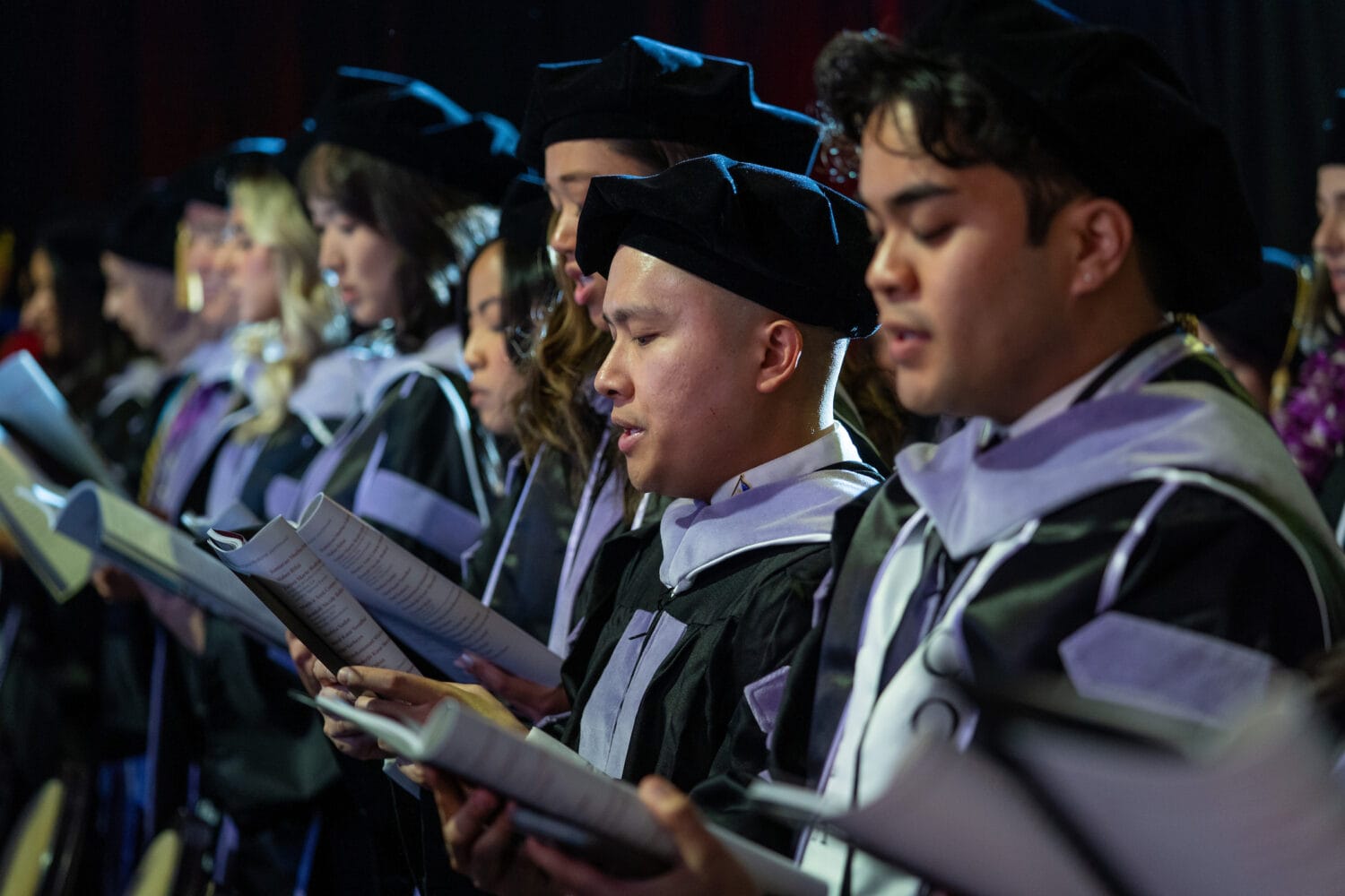 A group of graduates in caps and gowns stand in a row, holding programs and singing during a commencement ceremony.