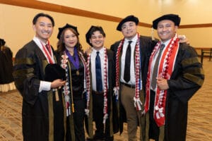Five people pose indoors; three wear graduation gowns, sashes, and caps, while two are in dress shirts and ties. All are smiling at the camera.