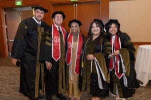 Five graduates in academic regalia and red stoles stand together indoors, smiling at the camera.