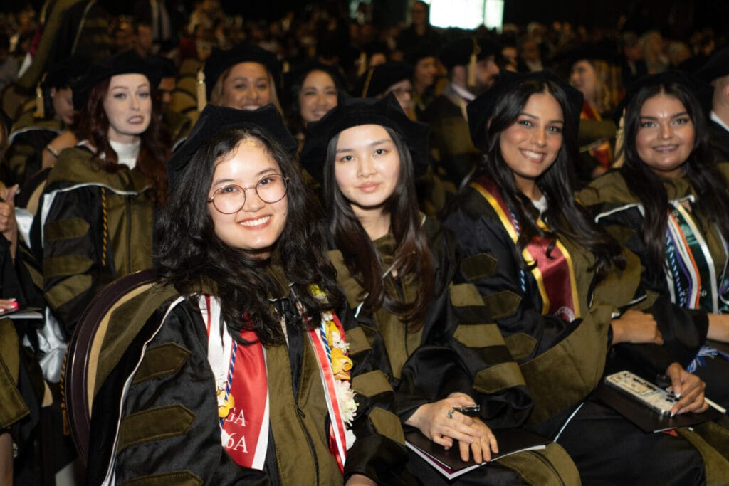 A group of graduates in caps and gowns sit together at a commencement ceremony, smiling and holding diplomas.