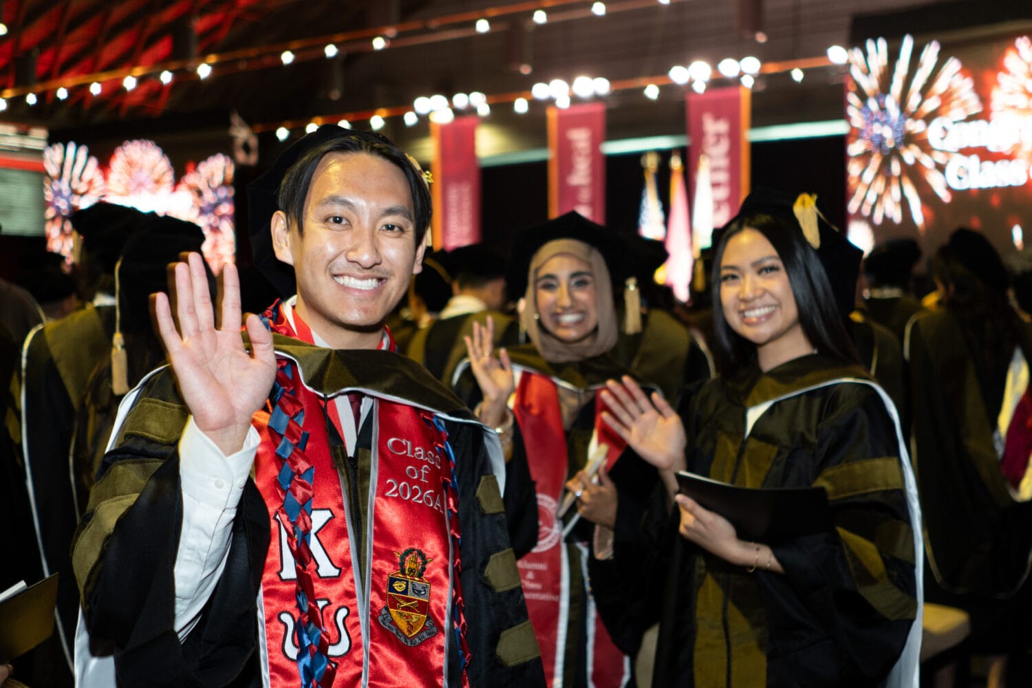 Three graduates in caps and gowns smile and wave at a commencement ceremony, with festive lights and banners in the background.