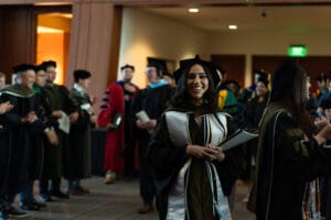 A graduate in cap and gown smiles and holds a diploma while standing indoors among other graduates and faculty members during a graduation ceremony.