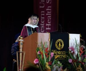 A woman in academic regalia speaks at a podium with a Western University of Health Sciences banner during a formal ceremony.