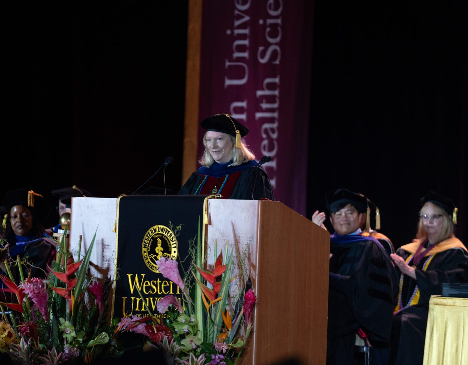 A person in academic regalia speaks at a podium during a graduation ceremony, with faculty members seated behind and floral arrangements in front.