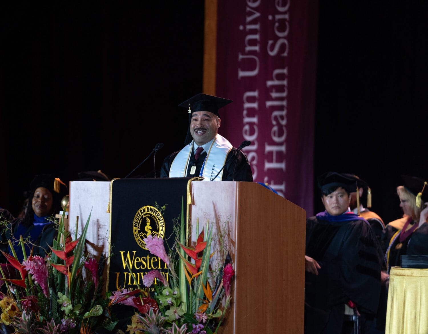 A graduate in a cap and gown speaks at a podium during a commencement ceremony at Western University of Health Sciences, with faculty seated behind him.