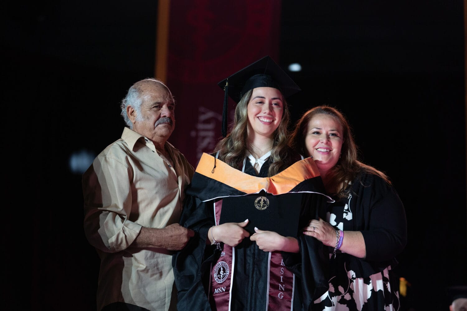 A graduate in cap and gown stands smiling between two adults, likely her family members, during a graduation ceremony.