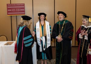 A graduate in cap and gown stands smiling with three university officials in academic regalia at a Western University of Health Sciences ceremony.