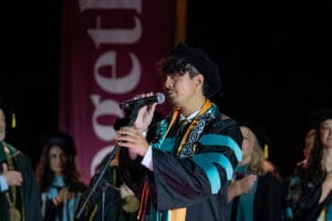 A graduate in academic regalia speaks or sings into a microphone on stage during a ceremony, with others in similar attire in the background.