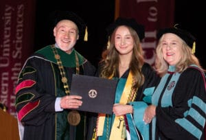 Three people in academic regalia pose on stage at a graduation ceremony. The person in the center holds a diploma and is flanked by two university officials.