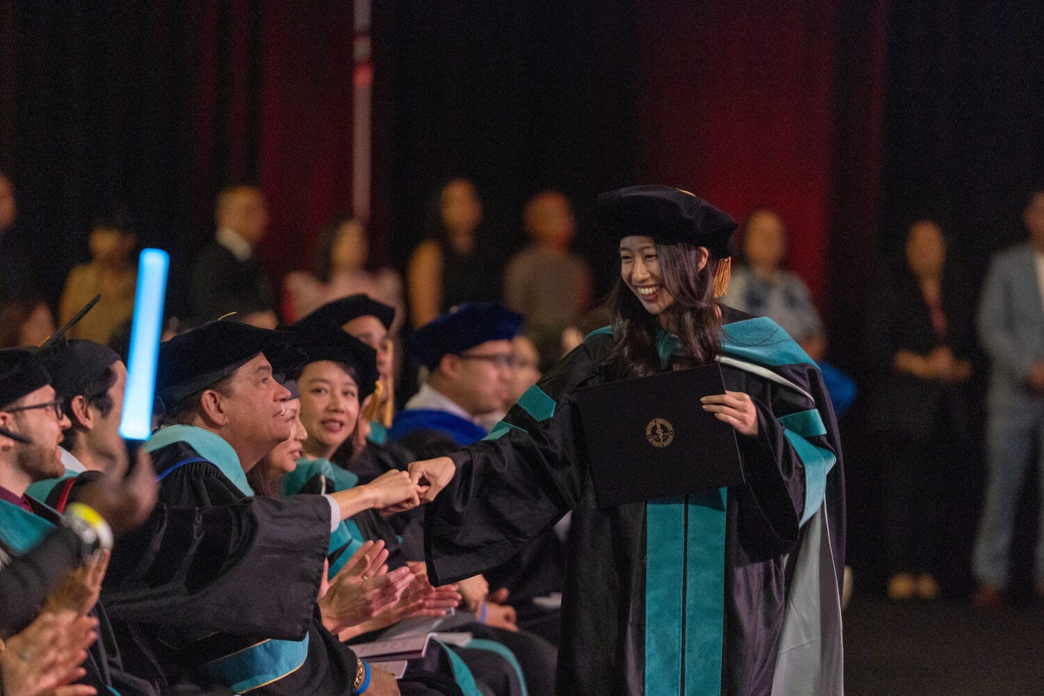 A graduate in academic regalia smiles and fist-bumps a seated faculty member while holding her diploma at a graduation ceremony.