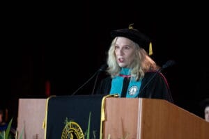 A woman in academic regalia speaks at a podium during a graduation ceremony. The podium displays a university seal.
