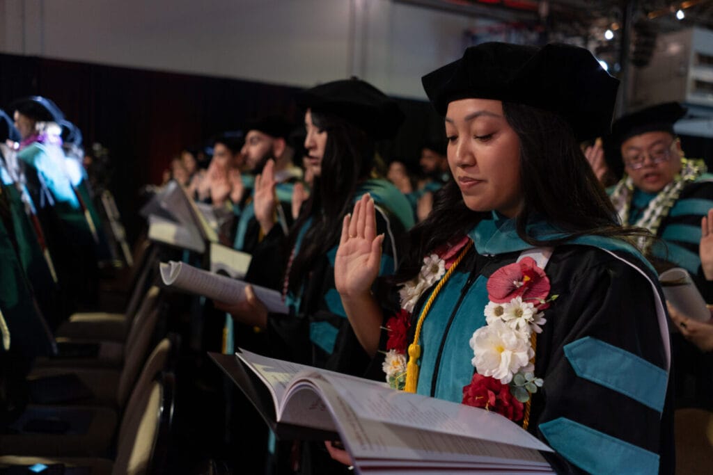 Graduates in academic regalia stand and hold up their right hands while reading from a booklet during a commencement ceremony.