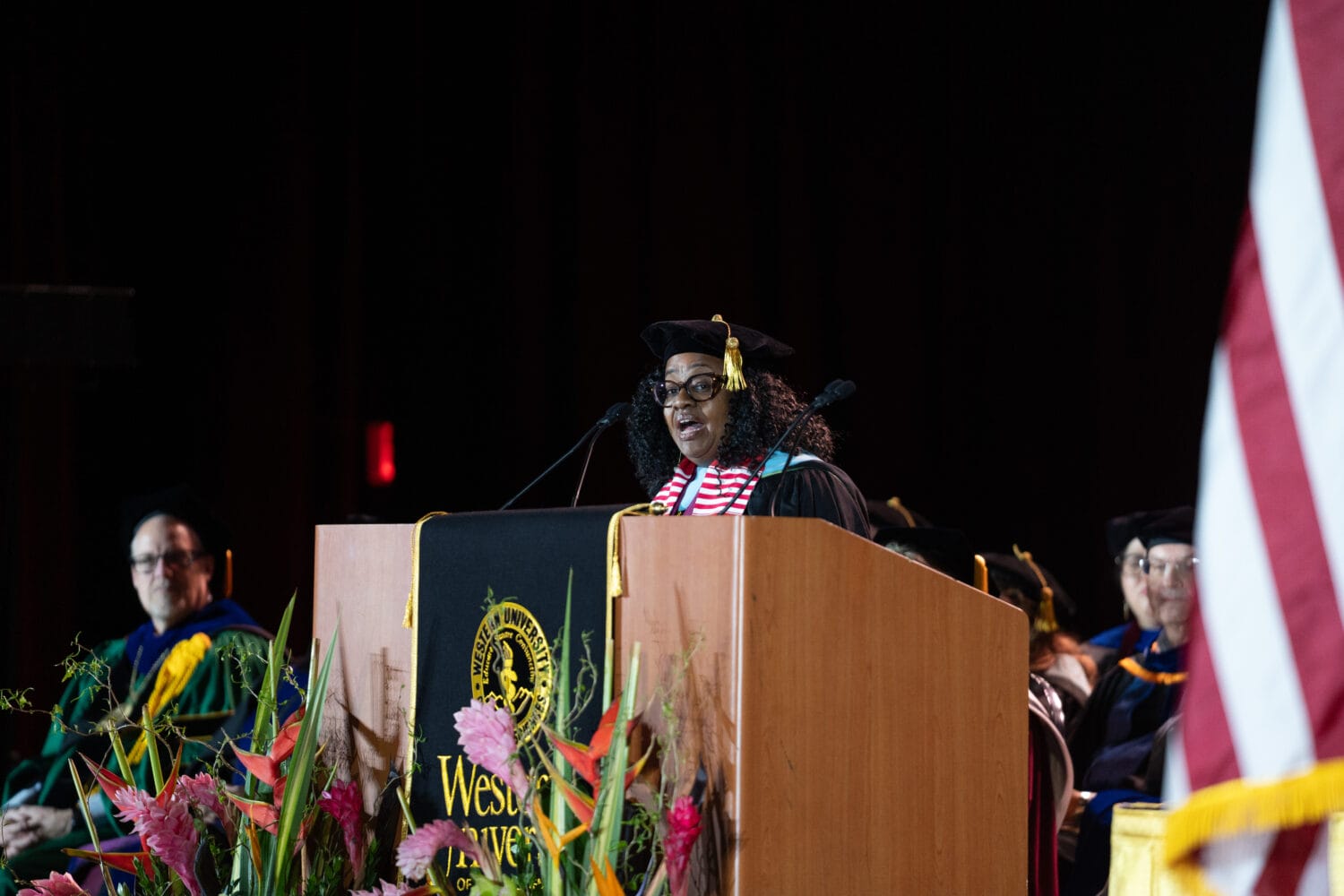 A woman in academic regalia speaks at a podium during a graduation ceremony.