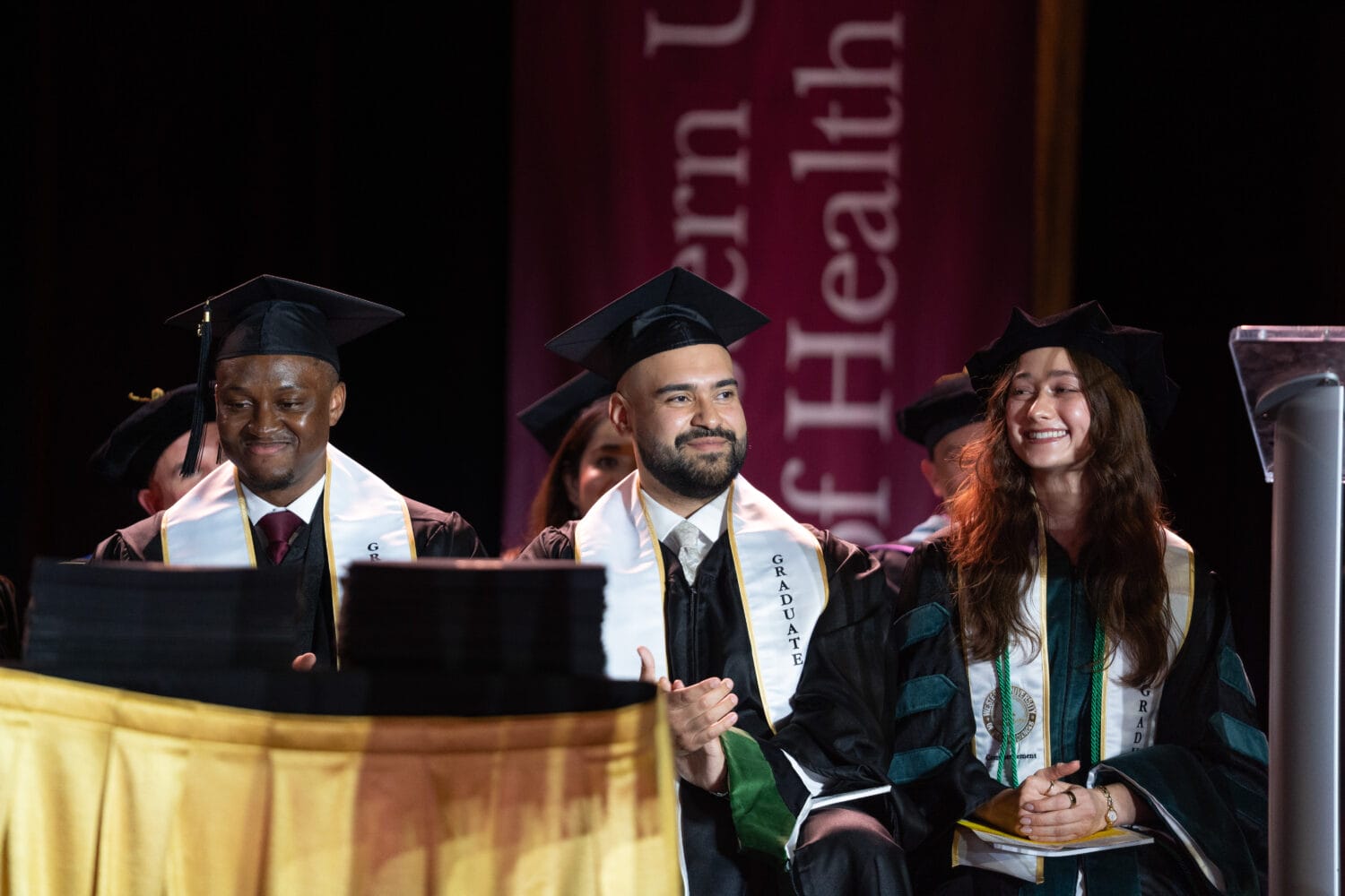 Three graduates in caps and gowns sit on stage, smiling and clapping during a commencement ceremony. A podium and university banner are visible in the background.