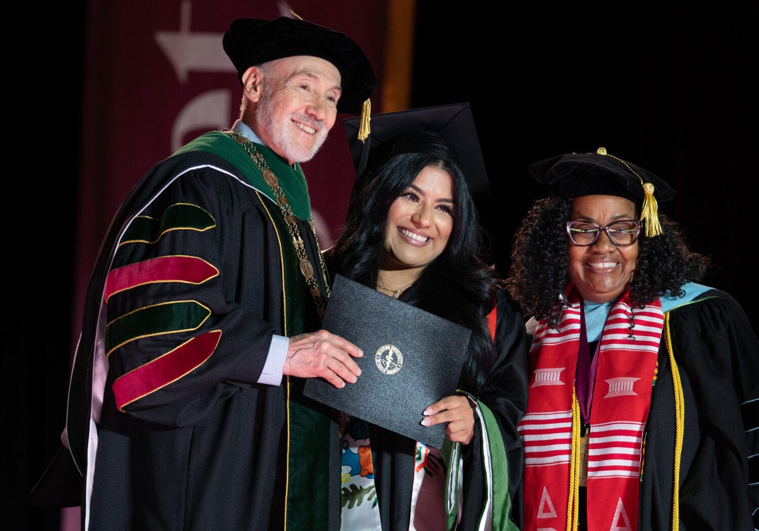 A graduate in cap and gown holds a diploma while standing between two officials in academic regalia during a commencement ceremony.
