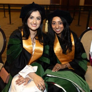 Two women wearing academic regalia and gold stoles sit next to each other, smiling at the camera during a graduation ceremony.