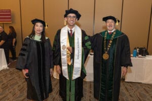 Three people in academic regalia pose indoors at a graduation event; the center person wears a sash reading "USC OST Alumni.