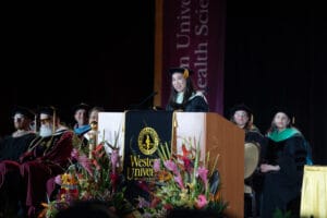 A speaker in academic regalia stands at a podium with "Western University of Health Sciences" signage during a graduation ceremony, with faculty seated behind and floral arrangements in front.