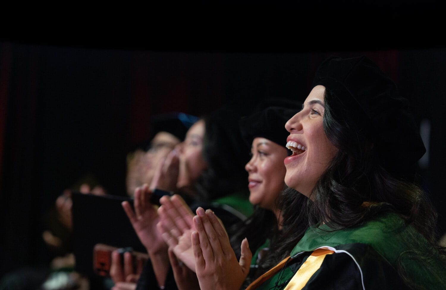 A group of graduates in caps and gowns sit and applaud, with one woman in the foreground smiling brightly.