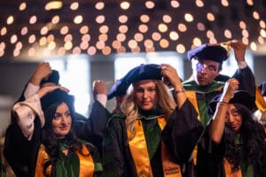 Four graduates in caps and gowns adjust their graduation caps during a ceremony, with blurred lights in the background.