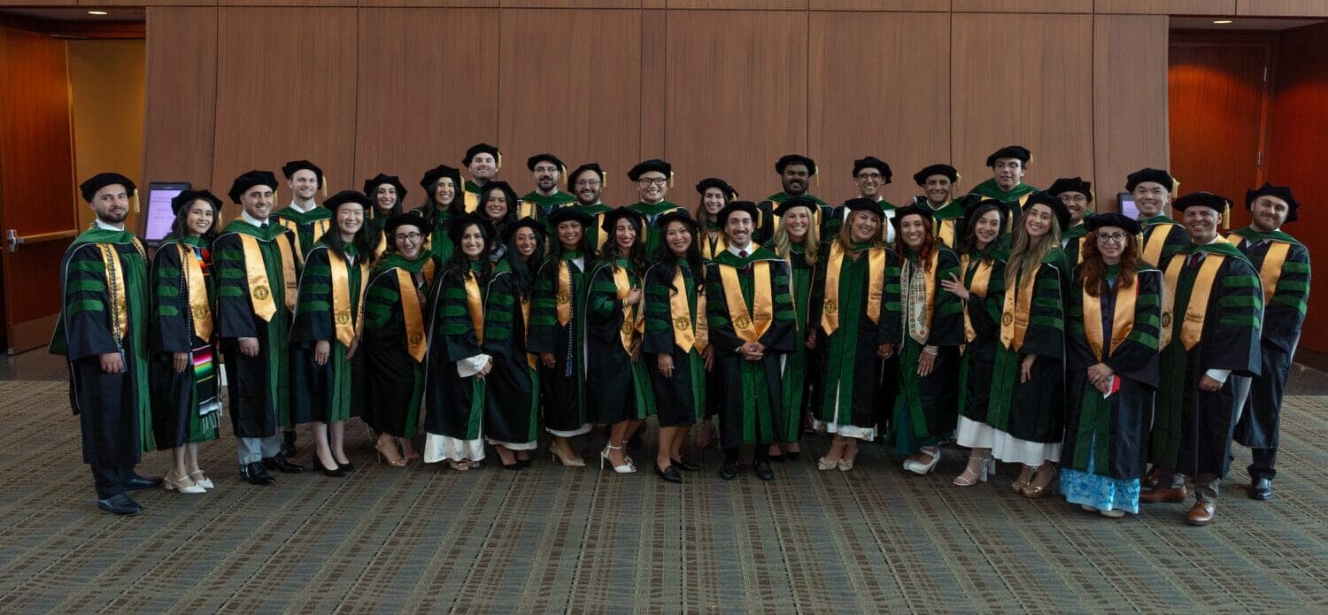 A group of graduates in caps and gowns pose in rows for a photo indoors against a wood-paneled wall.