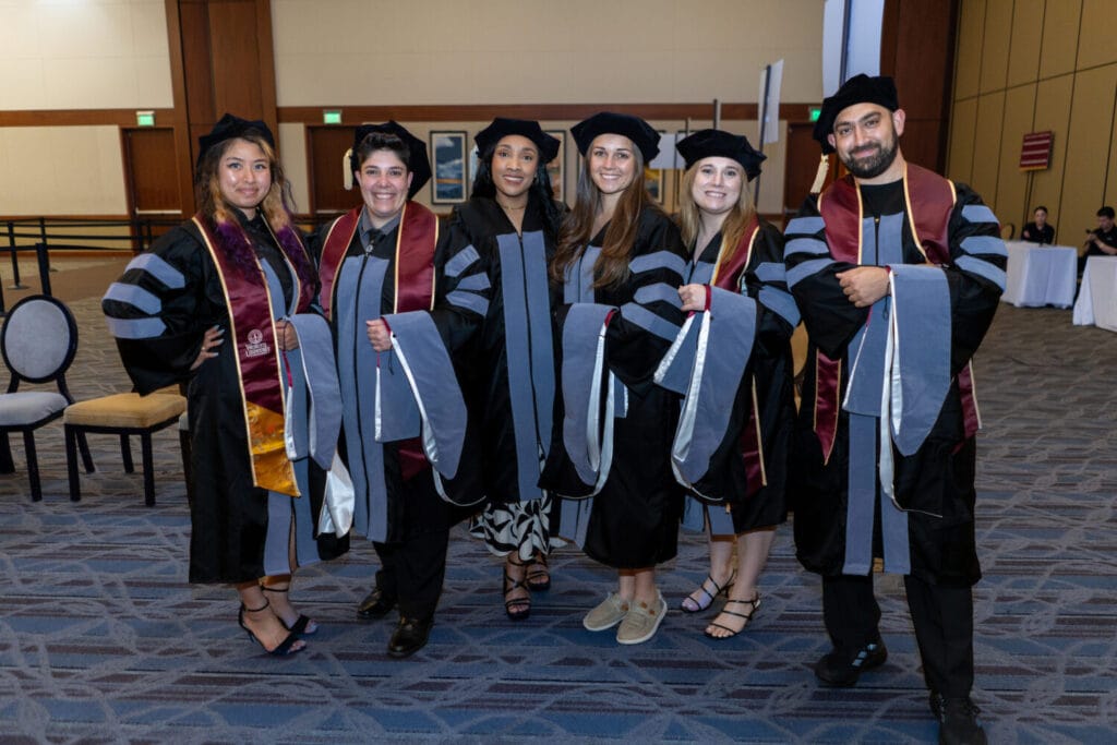 Six people in graduation regalia stand indoors, smiling at the camera. The group includes both men and women wearing black gowns and caps with grey and burgundy accents.