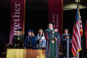 A group of university officials in academic regalia stand on stage at a graduation ceremony, with an American flag and banners in the background.
