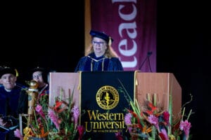 A speaker in academic regalia stands at a podium with the Western University of Health Sciences logo, delivering a speech at a graduation ceremony.