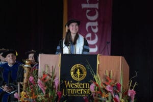 A graduate in academic regalia smiles at a podium during a Western University of Health Sciences ceremony, with faculty members seated behind her and floral arrangements in front.