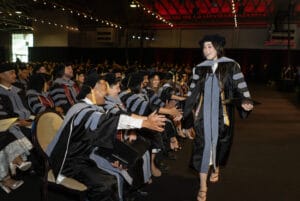 A graduate in cap and gown smiles and high-fives seated graduates during a commencement ceremony in an indoor venue.