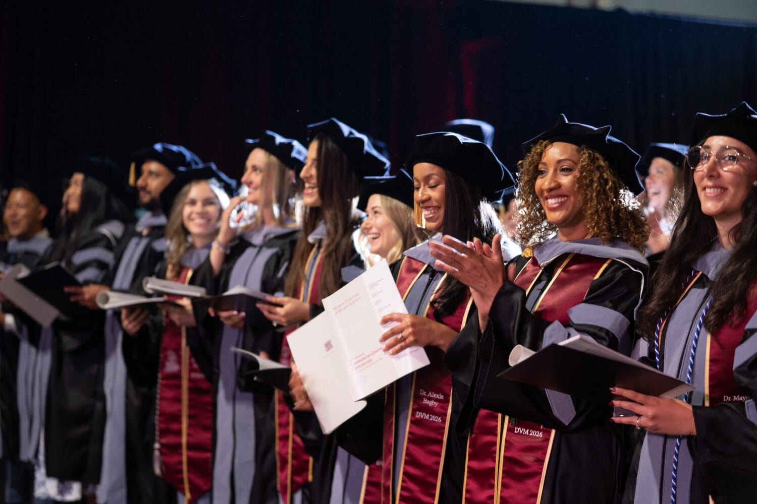 A group of graduates in academic regalia stand in a row, holding diplomas and smiling during a graduation ceremony.